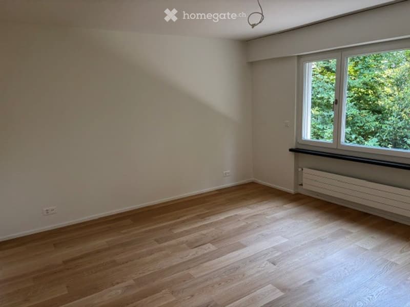 Empty room with wooden flooring, large window, radiator, and visible electrical outlets.