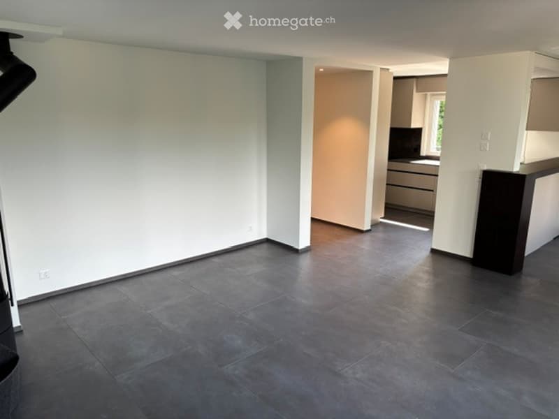 Empty room with dark grey tiled floor and white walls, connected to a modern kitchen area with grey cabinets and a countertop.