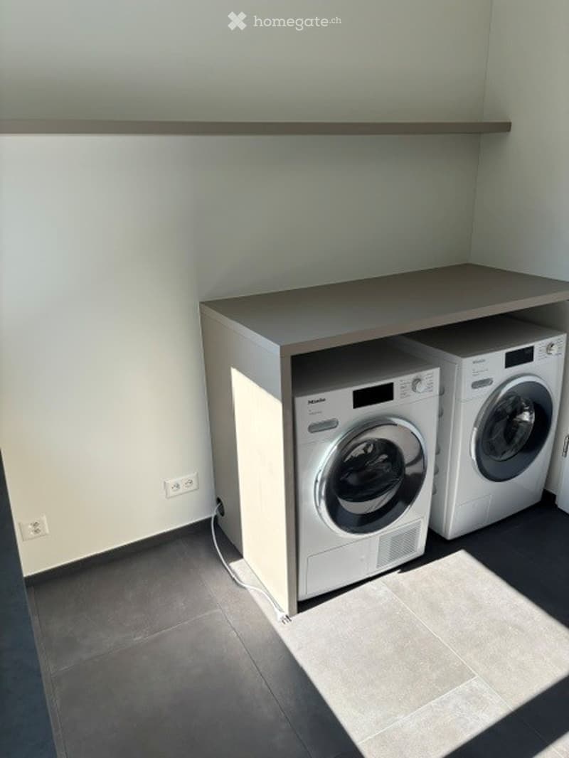 Modern, compact laundry area with a washing machine and dryer unit, white cabinetry, and tiled floor.