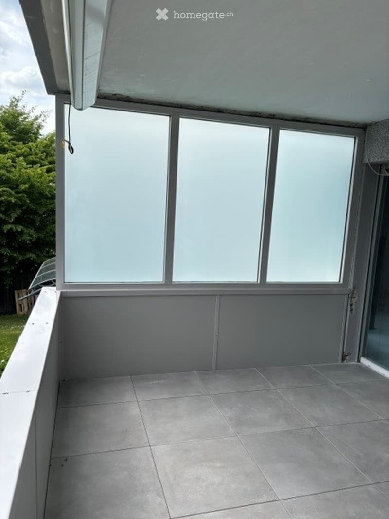 Empty balcony with tiled floor, large windows, and a view of greenery outside.