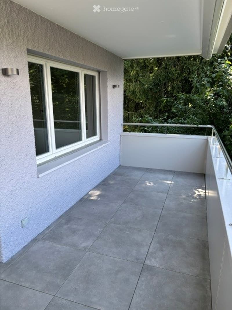 Modern balcony with tiled floor, white walls, large window offering a view of greenery, and a metal railing.