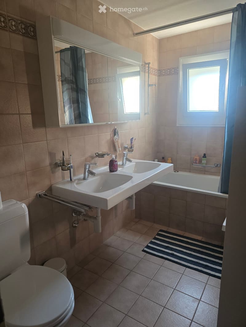 Modern bathroom with beige tiles, double sink, bathtub, toilet, and a large mirror above the sink. Includes a towel rack, soap dispenser, and toothbrush holder.
