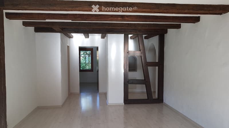 Empty hallway with wooden beams on the ceiling, white walls, and a wooden floor. A window at the end of the hallway allows natural light to enter.