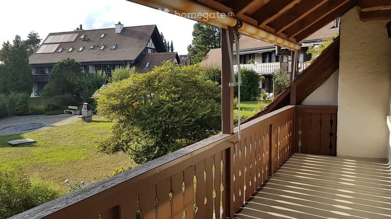 Balcony overlooking a well-maintained garden with trees, a bench, and a small structure. The railing is wooden with decorative cutouts, and the steps lead down to the garden.