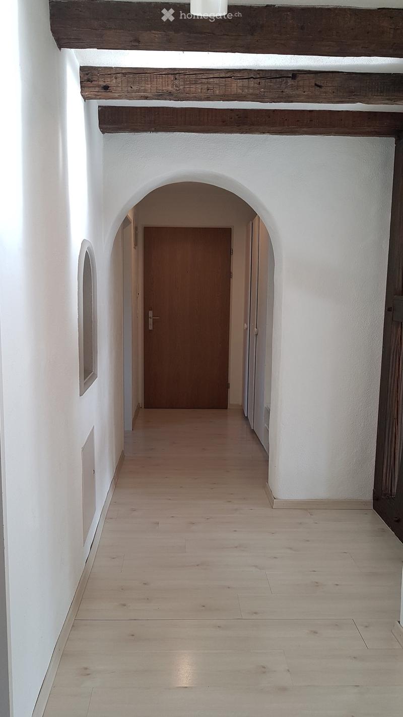 A long, arched hallway with white walls, wooden floor, and exposed wooden beams on the ceiling. A wooden door is visible at the end of the corridor.