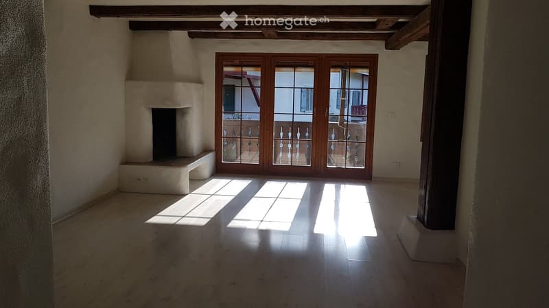 Empty room with wooden floor, exposed wooden beams, fireplace, and large glass doors leading to a balcony.