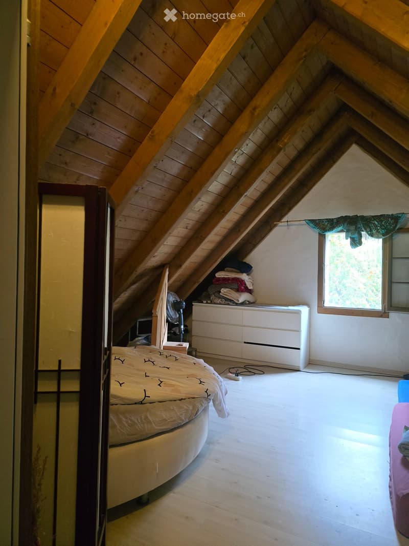 Cozy attic bedroom with wooden slanted ceilings, a round bed, a fan, a white drawer unit, and a window with a view.