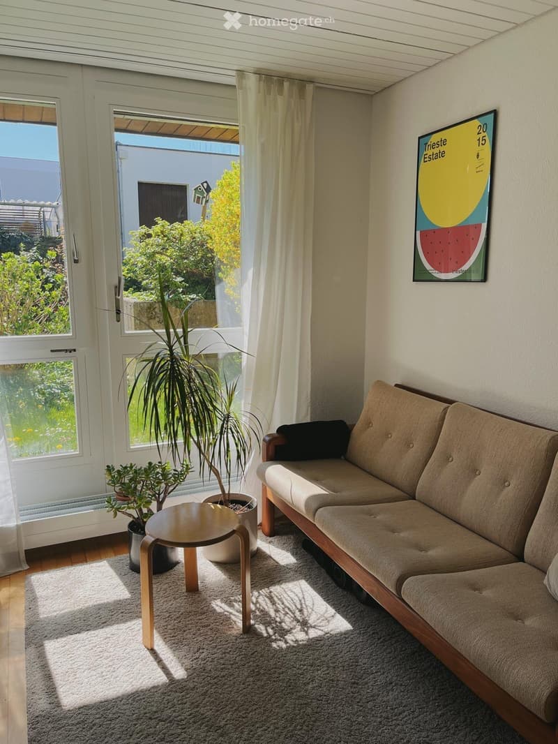 Bright living room with a beige sofa, small wooden table, potted plants, and a colorful poster on the wall. Large windows and a door lead to a garden view.