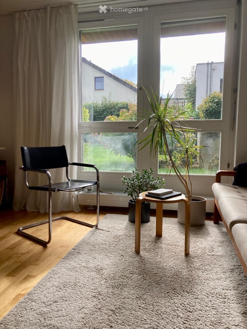 A modern living room with wooden flooring, a large window offering a view of the garden, a grey sofa, a black chair, a small wooden table with books, and potted plants.