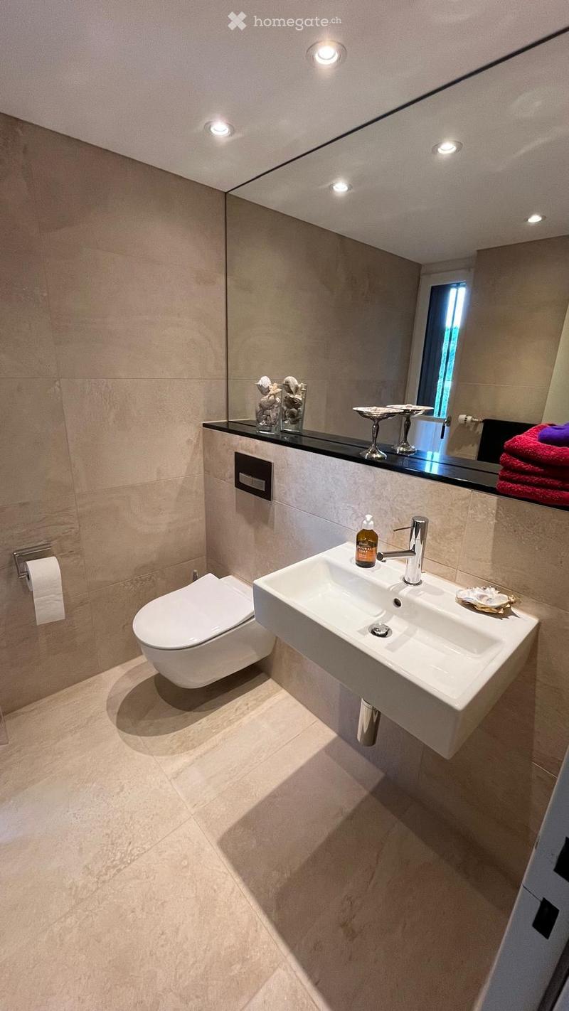 Modern bathroom with a white toilet, a white sink with a modern faucet, a large mirror, and decorative items on the counter. The walls are tiled in a light beige color, and the floor is also tiled in a matching light beige color.