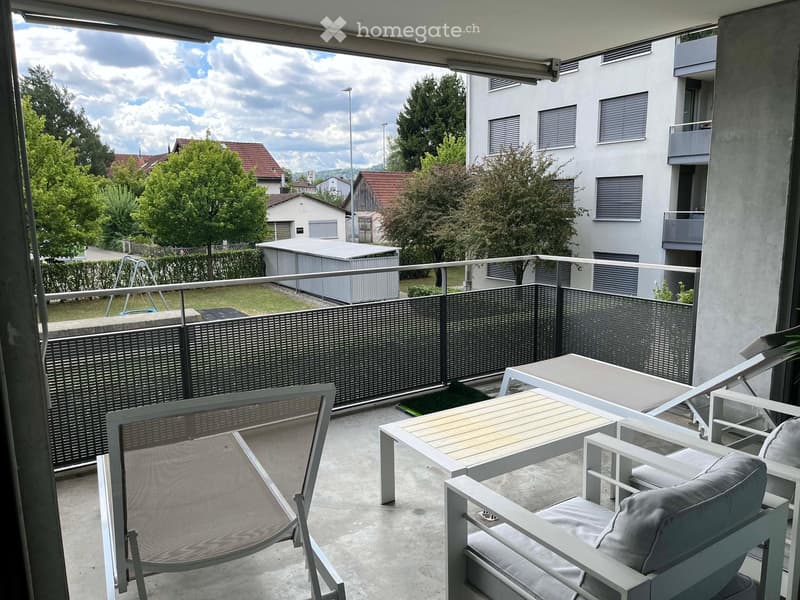 Balcony with a table and chairs, overlooking a residential area with trees and houses.