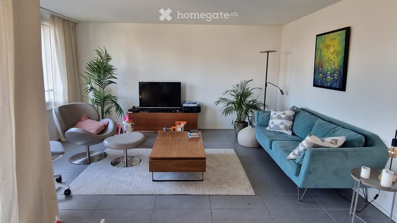 Modern living room with a grey sofa, wooden coffee table, grey chair, and television on a wooden stand. The room features large windows with curtains, a floor lamp, and decorative plants.