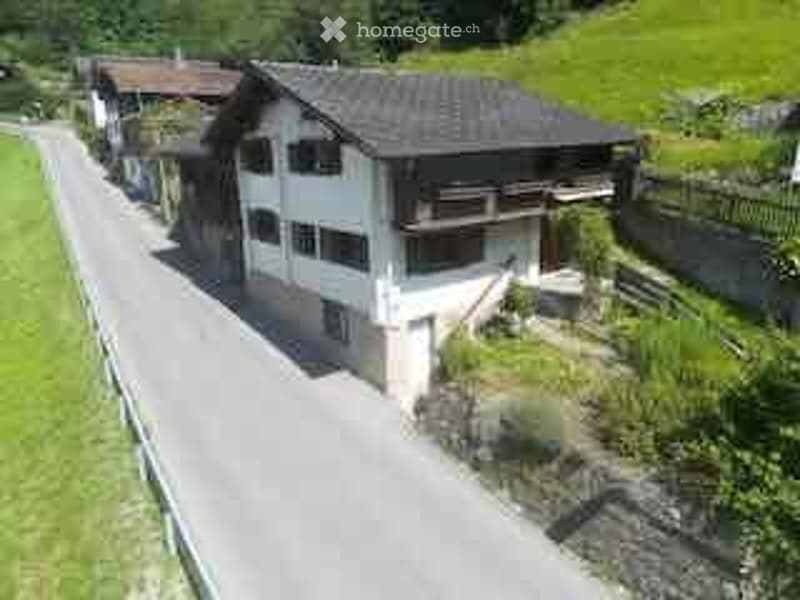 A multi-story house with a dark roof and white walls, situated on a hillside with a driveway leading to it. The house has multiple windows and a balcony on the upper level.