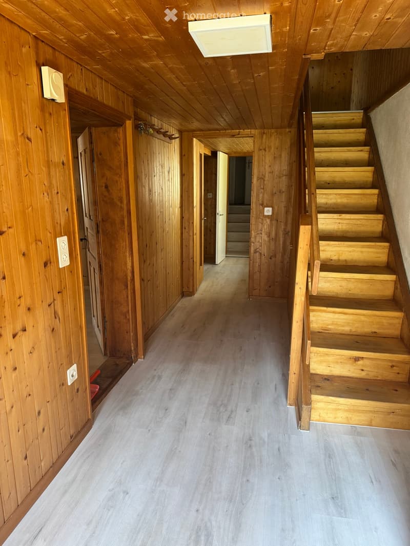 Wooden-paneled hallway with a staircase leading upwards, featuring light-colored wooden steps and railings, and several doors along the walls.