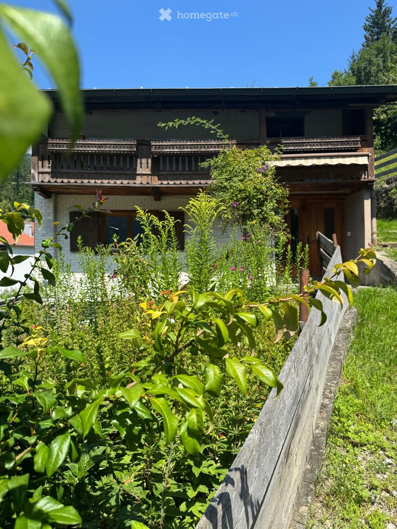A garden area with lush green plants and a wooden fence, leading to a two-story house with a balcony and wooden detailing.