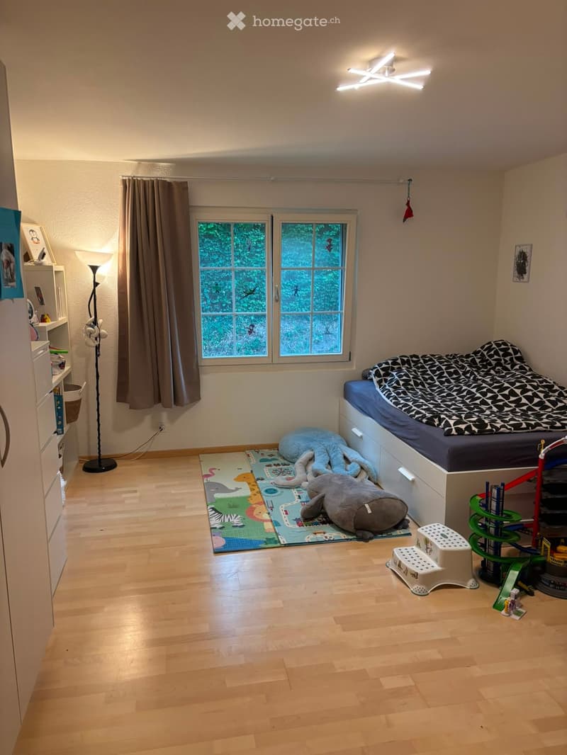 A well-lit bedroom with a bed, a colorful play mat, stuffed animals, a small stool, and a toy car. The room features wooden flooring, a large window with curtains, and a bookshelf with various items.