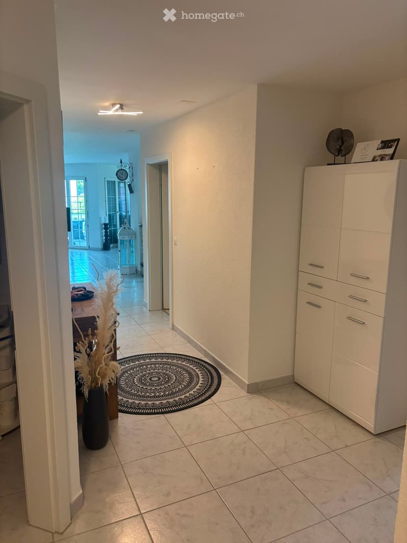 Hallway with tiled floor, white walls, built-in white cabinet with drawers, decorative black and white round rug, vase with dried plants, and a view into another room with a clock and lantern.