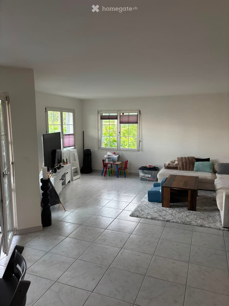 Bright living room with tiled flooring, large windows, a sofa with cushions, a coffee table, a small table with chairs, and a TV stand.