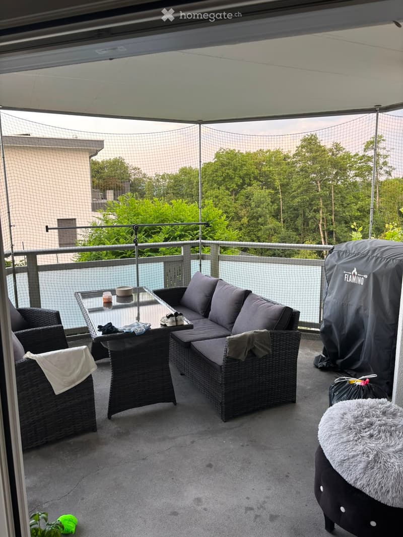 Balcony with a wicker sofa, glass table, wicker chairs, and a fur-covered stool, featuring a covered grill and a view of greenery.