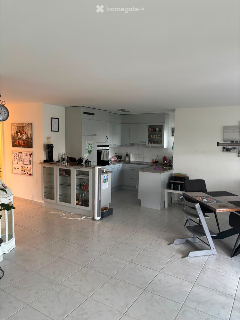 Modern kitchen with grey cabinets, tiled floor, and a dining area with a table and chairs. Appliances include a refrigerator, oven, coffee maker, and a fan. There are also various kitchen items on the counter and a picture frame on the wall.