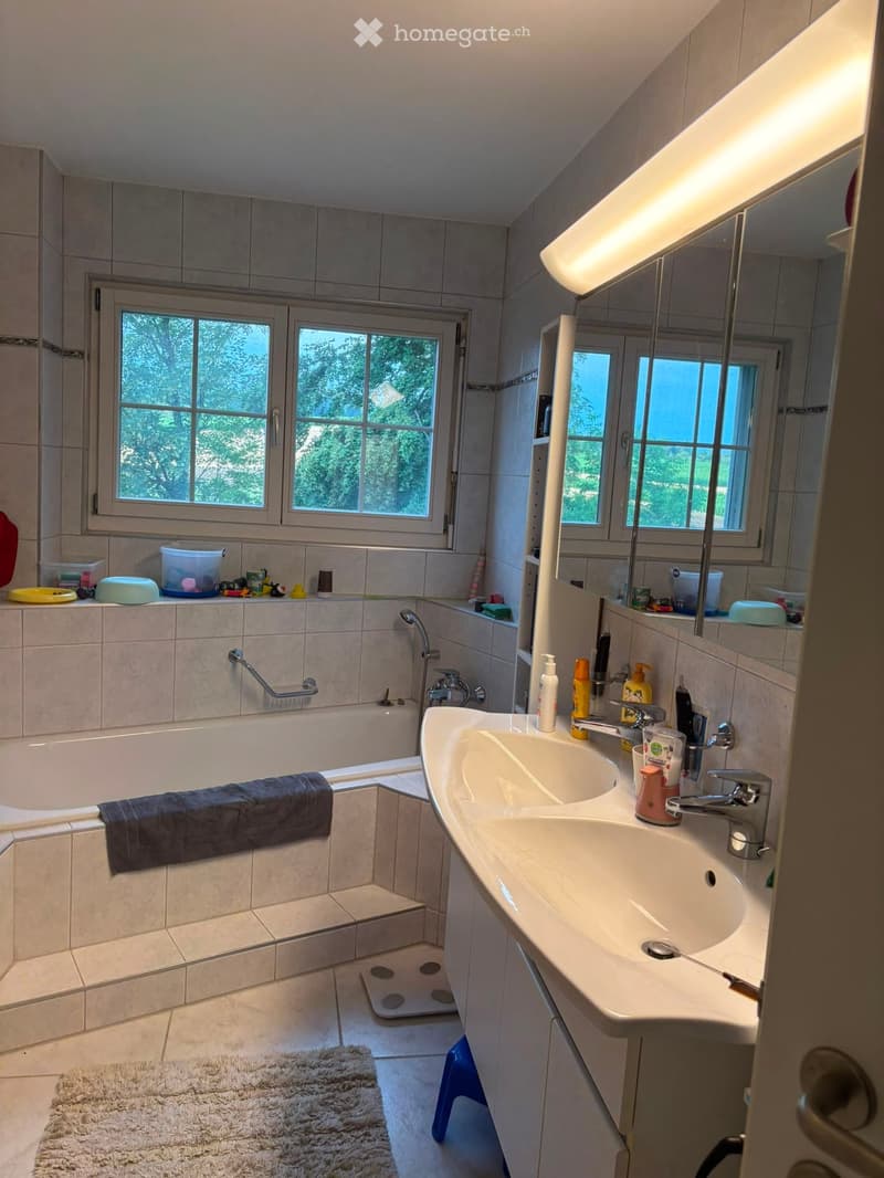 Bathroom with a bathtub, sink, mirror, and various toiletries. The room has tiled walls and a window letting in natural light.