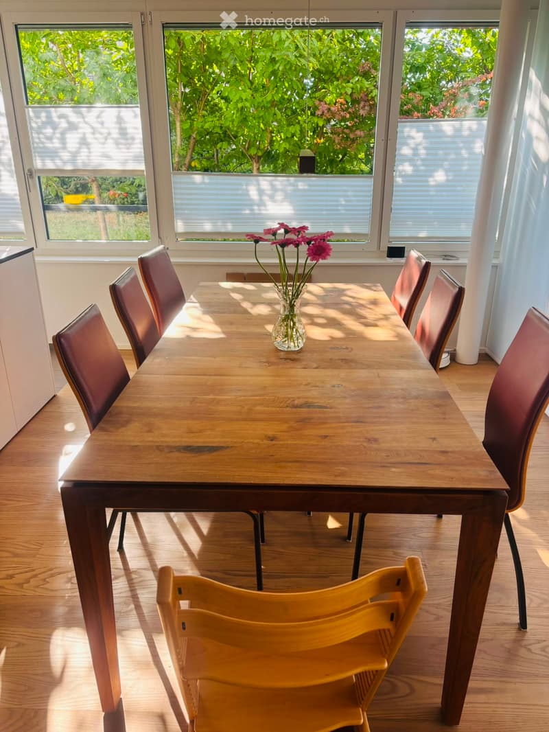 Bright dining area with a wooden table, six chairs, and a vase of pink flowers on the table. Large windows with blinds allow natural light to fill the space.