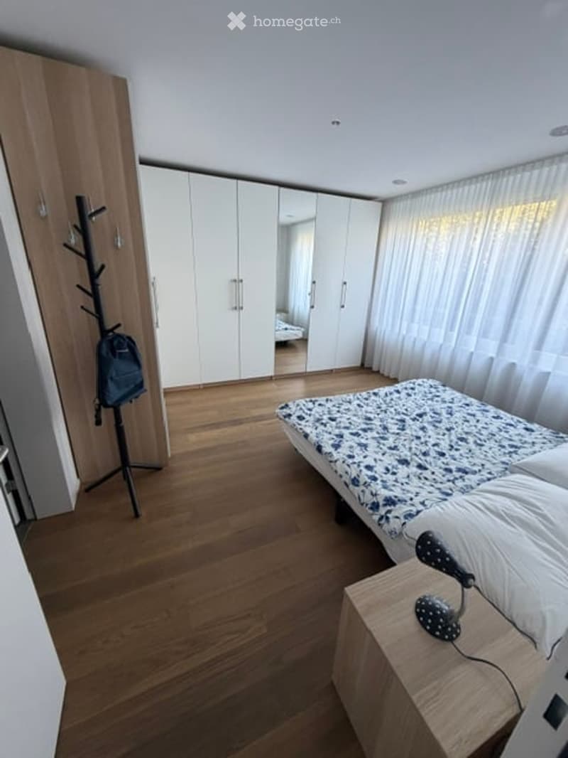 Modern bedroom with wooden flooring, a bed with blue and white patterned bedding, a nightstand with a lamp, a large white wardrobe, and a coat rack with a backpack.