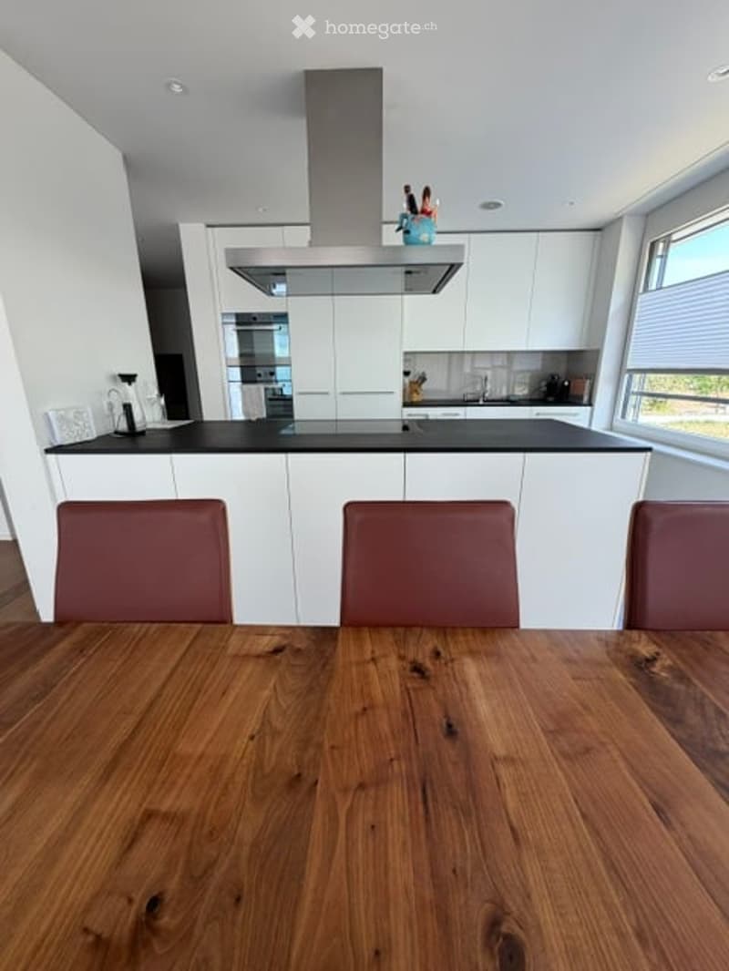 Modern kitchen with white cabinets, black countertops, a wooden dining table with three red chairs, and a kitchen island with a coffee maker and range hood.