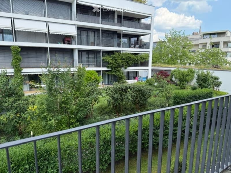 Balcony overlooking a multi-story residential building with several balconies, greenery, and a view of neighboring structures.