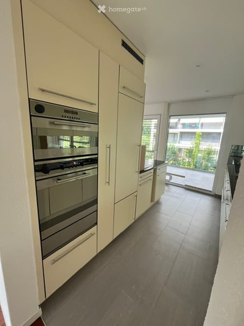 Modern kitchen with light-colored cabinets, stainless steel oven, and large window offering a view of greenery.