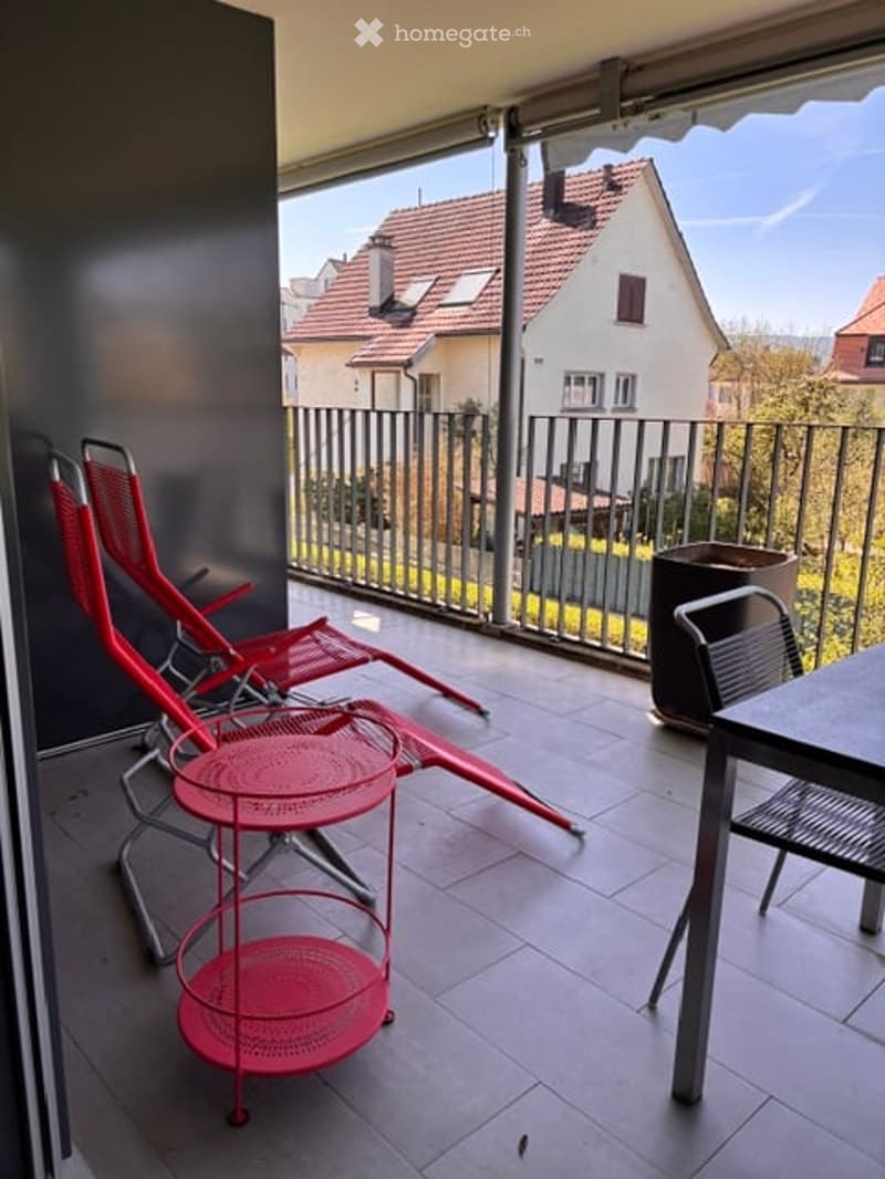 Balcony with red chairs, small round table, and a view of neighboring houses.