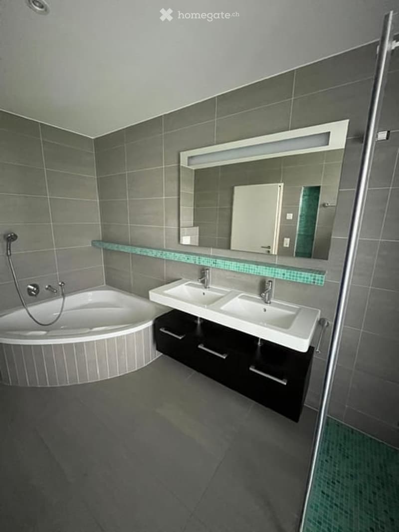 Modern bathroom with a sleek bathtub, double sink vanity, large mirror with integrated lighting, and dark grey tiles.