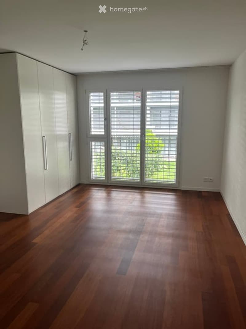 Empty room with wooden flooring, white walls, large windows with blinds, and white built-in wardrobes.