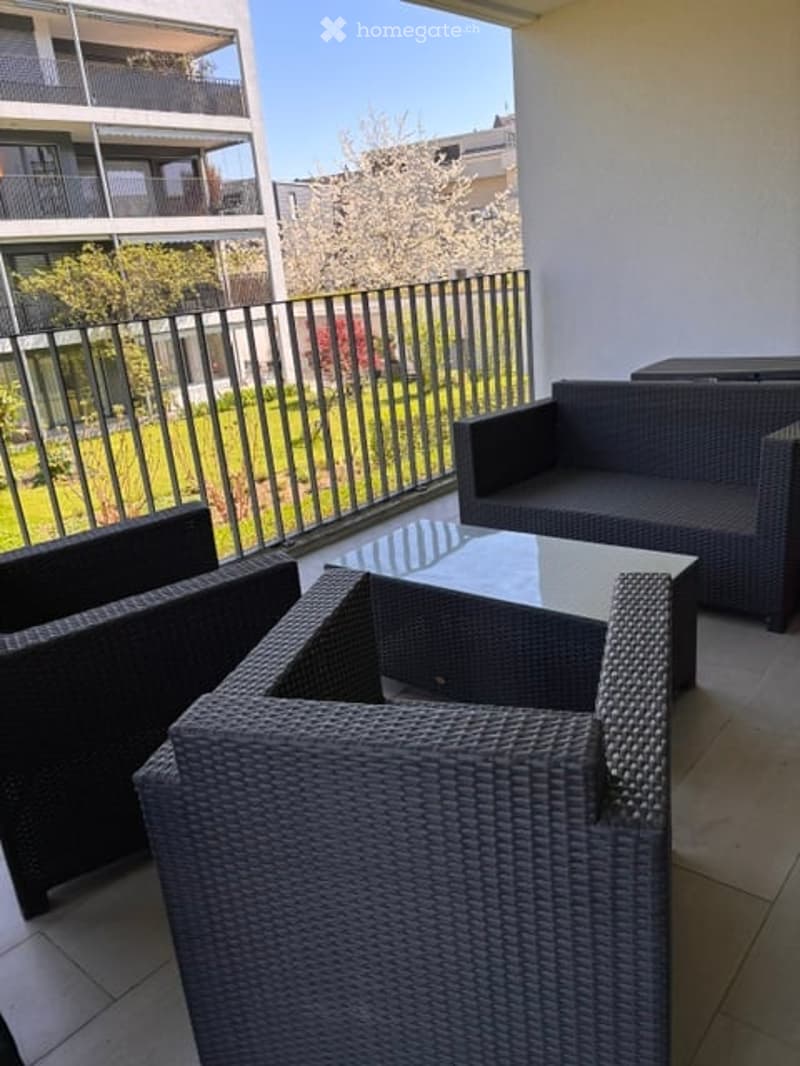 Balcony with wicker furniture including a sofa, chairs, and a coffee table, overlooking a green area with trees and neighboring buildings.