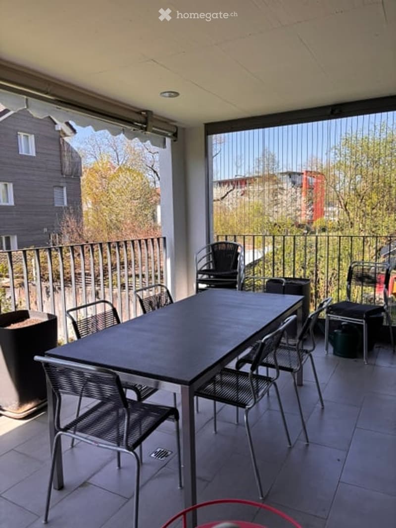 Balcony with a black dining table and chairs, overlooking a cityscape with trees and buildings.