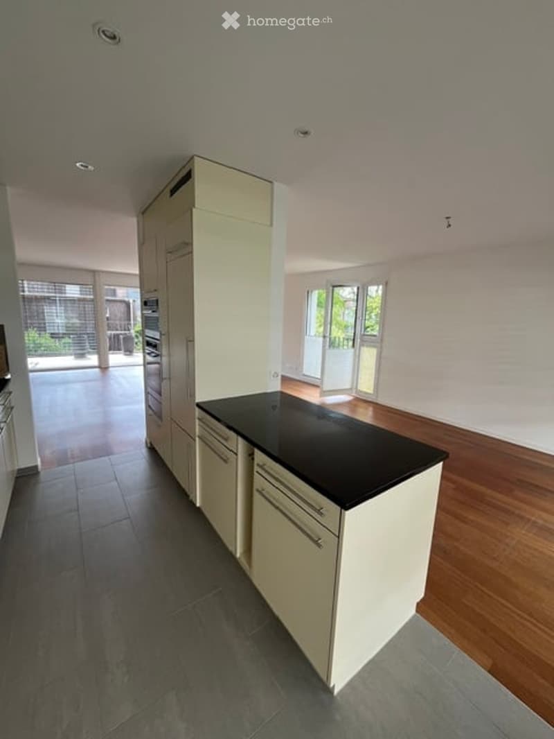 Modern kitchen with white cabinets, black countertop, and built-in oven. The space features a mix of dark and light flooring, with large windows providing natural light and access to an outdoor area.
