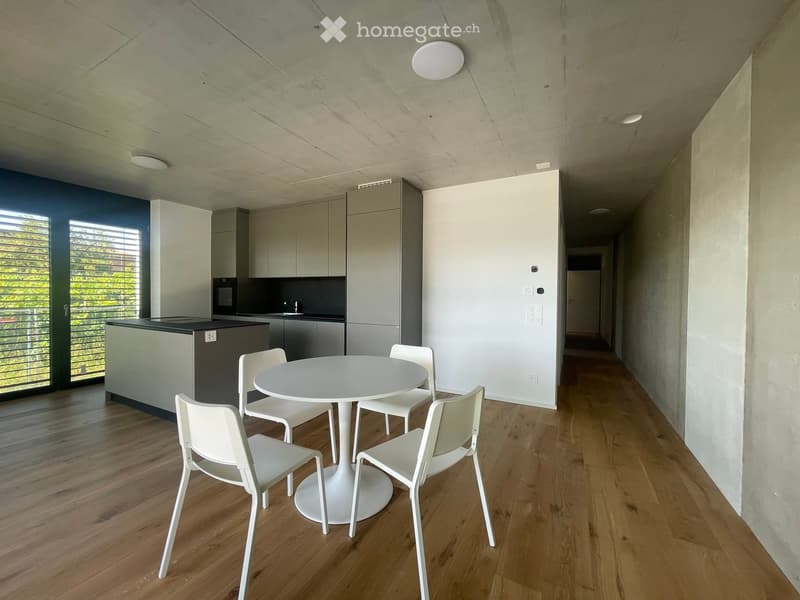 Modern, minimalist dining area with a white round table and four white chairs, adjacent to a sleek kitchen with grey cabinets and a black countertop.
