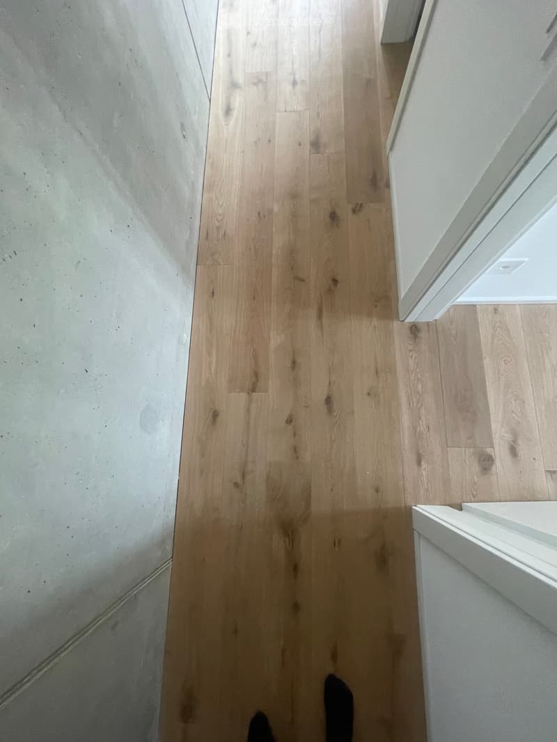 A narrow hallway with light wooden flooring and white walls, featuring a white baseboard and a visible door frame on the right side.