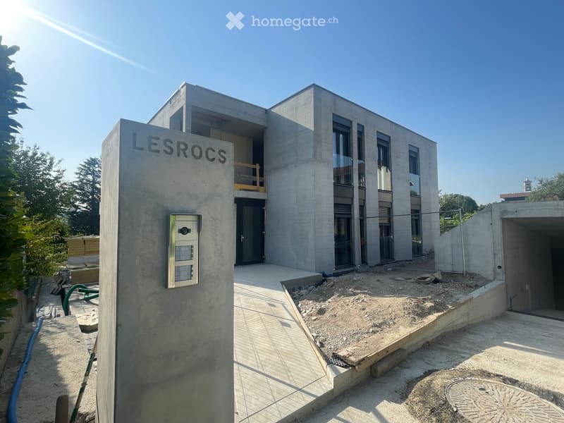 Modern two-story concrete building with large windows and a raised ground floor, featuring a security intercom and a partially constructed entrance area.