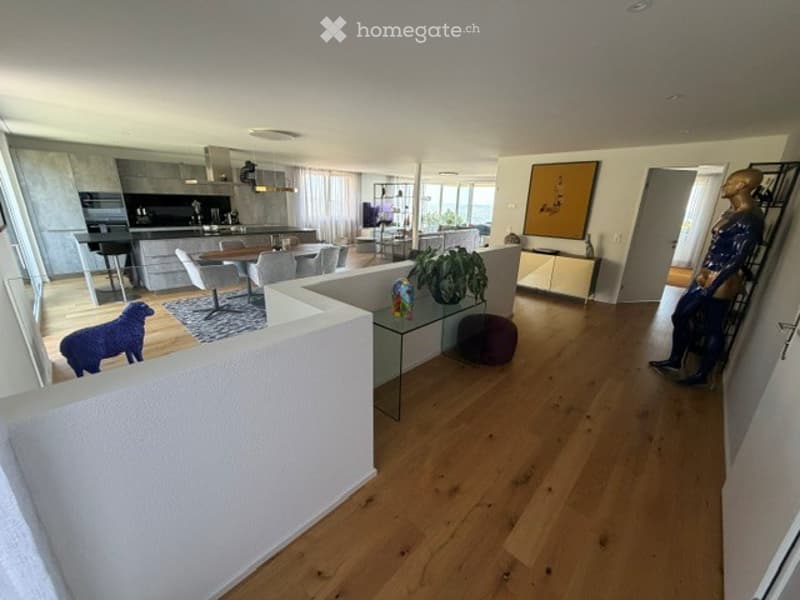 Modern open-plan living and dining area with light wood flooring, white walls, and contemporary furniture including a sofa, coffee table, dining table, and chairs. A small kitchen area is visible in the background with a kitchen island and bar stools.