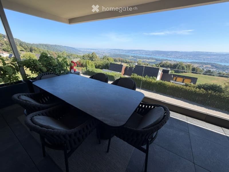 Balcony with a panoramic view of a valley and lake, featuring a table and chairs.
