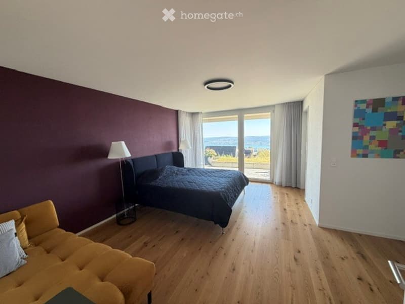 Bright bedroom with wooden flooring, large windows offering a sea view, a dark-colored bed, a mustard-colored sofa, and two lamps.