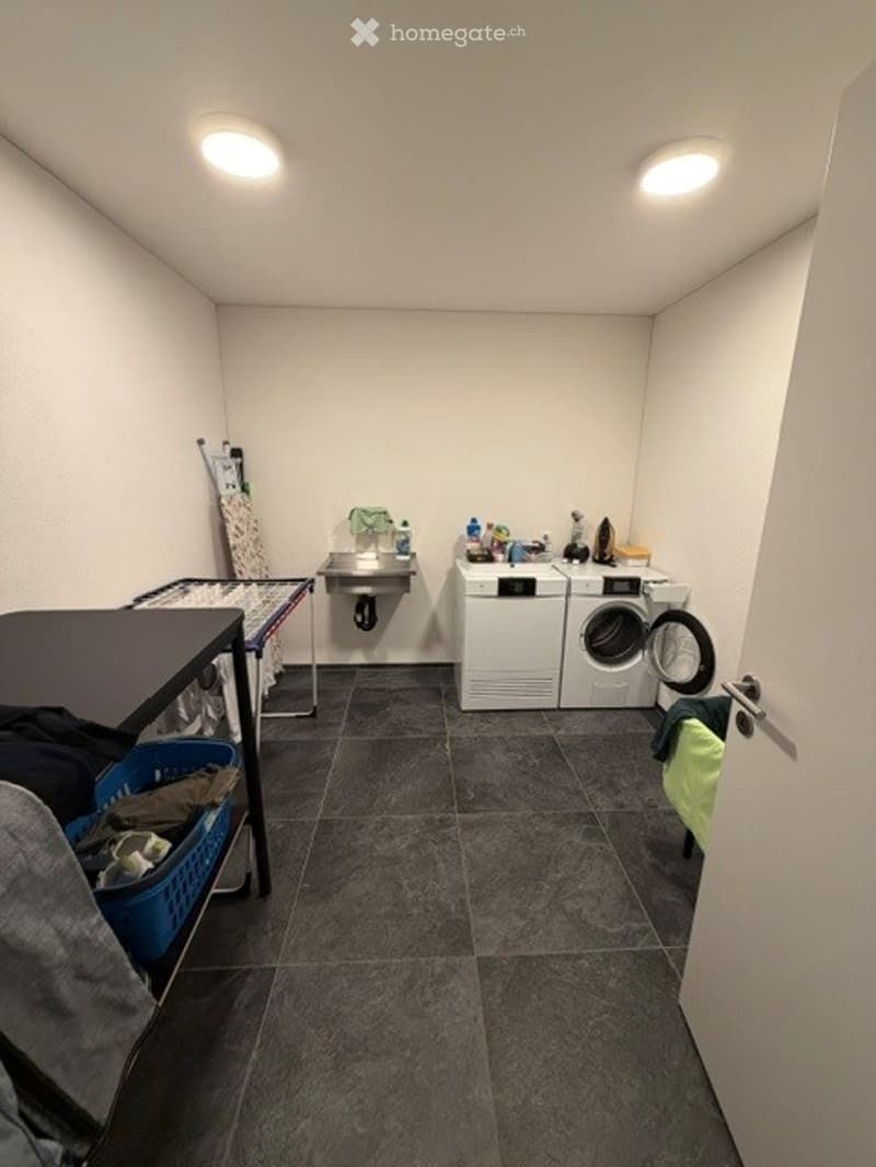 Laundry room with a washing machine, dryer, small table, chair, and a sink. The room has a tiled floor and white walls.
