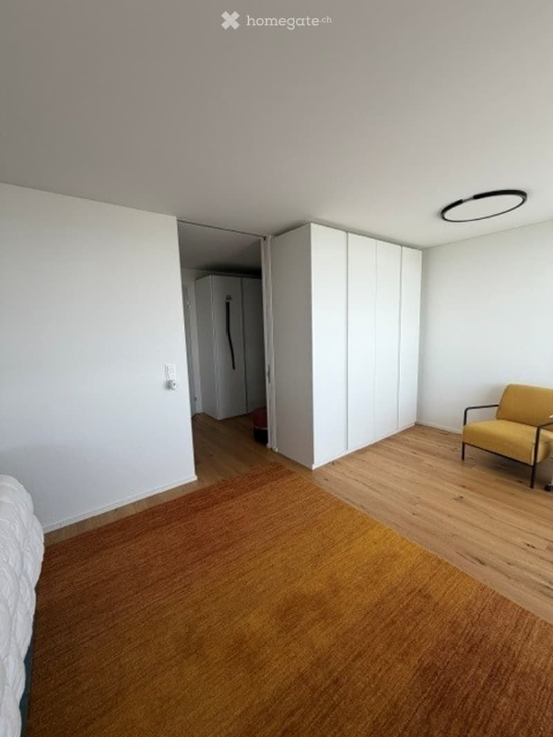 Bright, minimalist bedroom with wooden flooring, white walls, a yellow sofa, a large wardrobe, and a bed with white bedding.