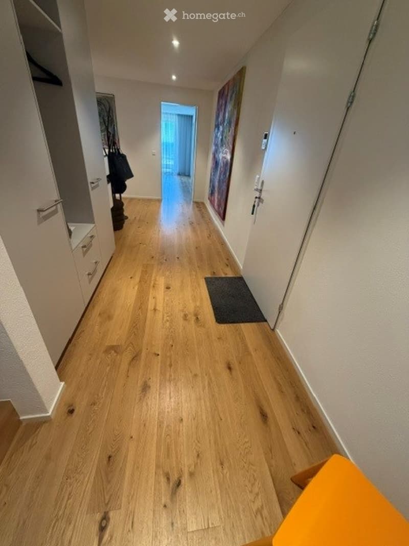 A well-lit hallway with light wooden flooring, white walls, and a painting on one side. A small black rug is placed near the center, and a door at the end leads to another room.