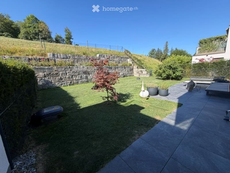 Well-maintained garden with artificial grass, stone retaining wall, small tree, potted plants, and patio area with seating.
