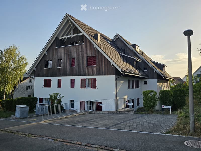 Two-story house with a combination of wooden and white exterior walls, red shutters, and a sloped roof. The house features a fenced entrance area with a paved driveway.