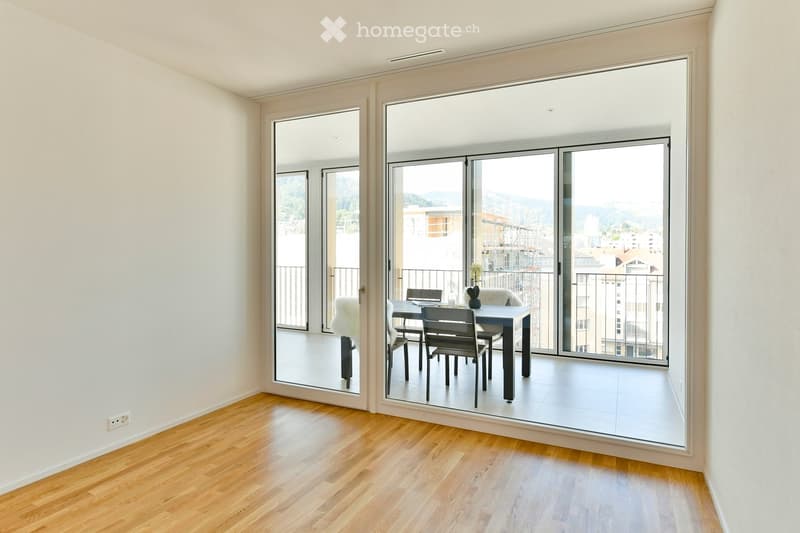 Bright room with wooden flooring, large sliding glass doors leading to a balcony, a dining table with chairs, and a laptop on the table.