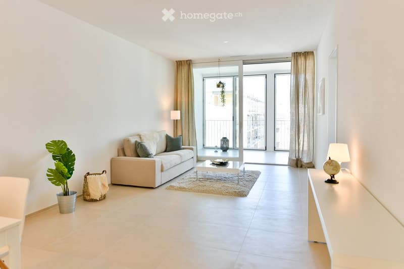 Bright living room with large sliding glass doors leading to a balcony, a white sofa with throw pillows, a coffee table on a small rug, a floor lamp, a table lamp, a globe, a potted plant, and a woven basket with a blanket.