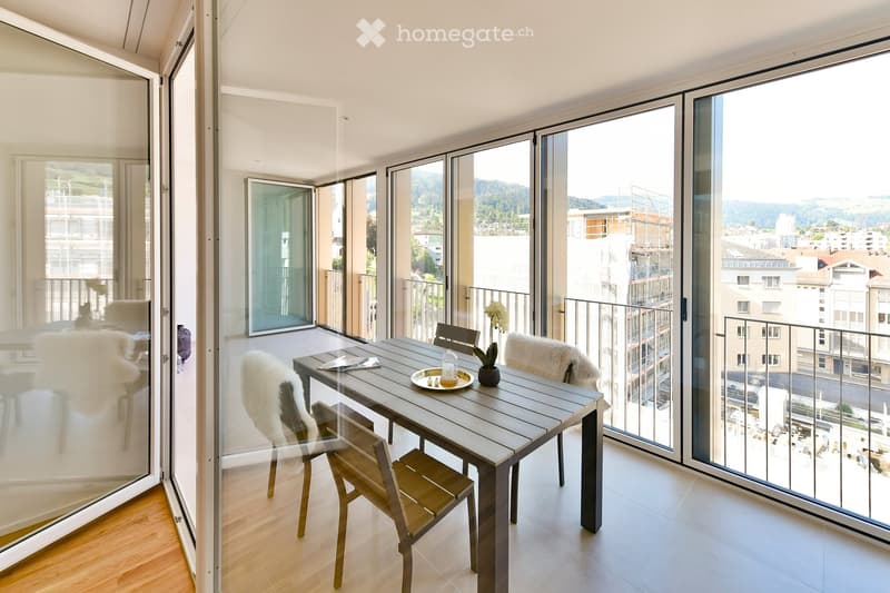 Modern dining area with a wooden table, four chairs, a vase with flowers, and a decorative plate on the table. Large glass windows offer a view of the city and mountains.